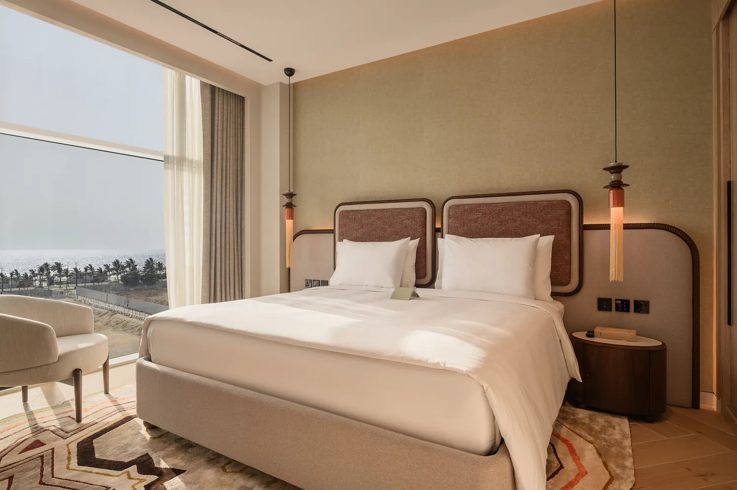 Deluxe Suite: a crisp white bed beside floor-to-ceiling windows looking over palms to the Red Sea.