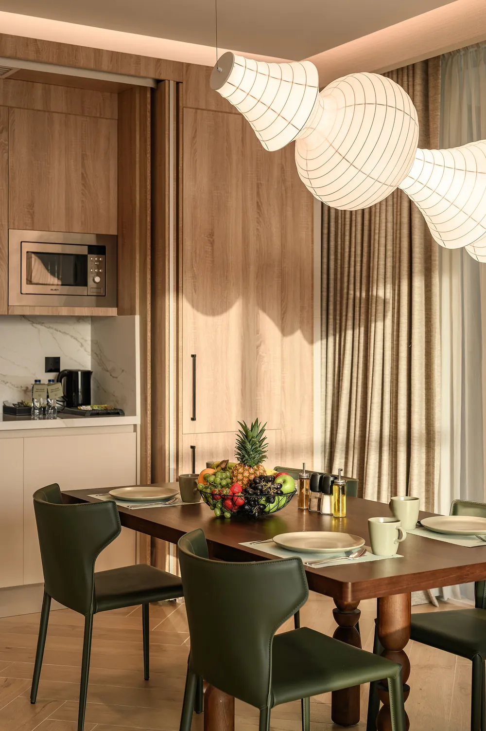 In-suite dining: a walnut table set for breakfast with fresh fruit beneath paper lanterns, beside an oak kitchenette.